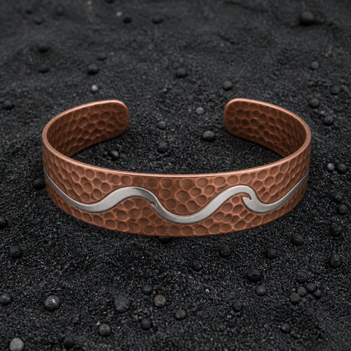 Early concept of a hammered cuff with a wave running across it, on black sand