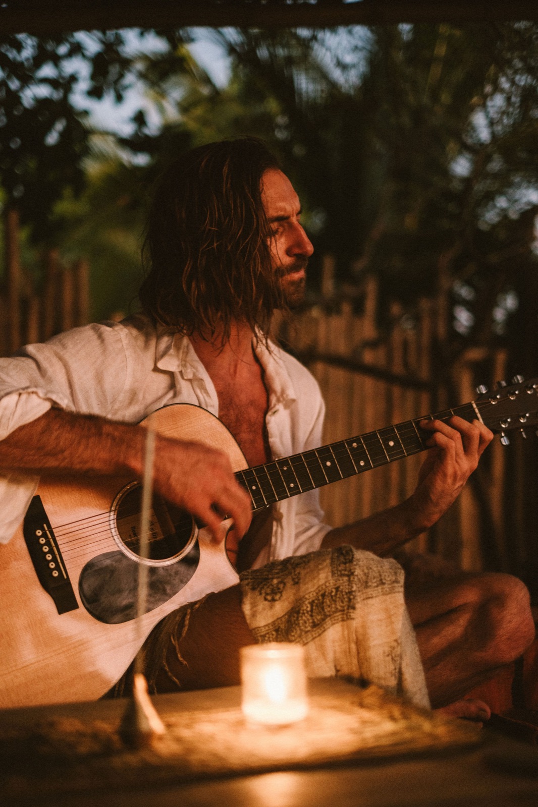 Enrique plays guitar by candlelight in an open bamboo room at dusk